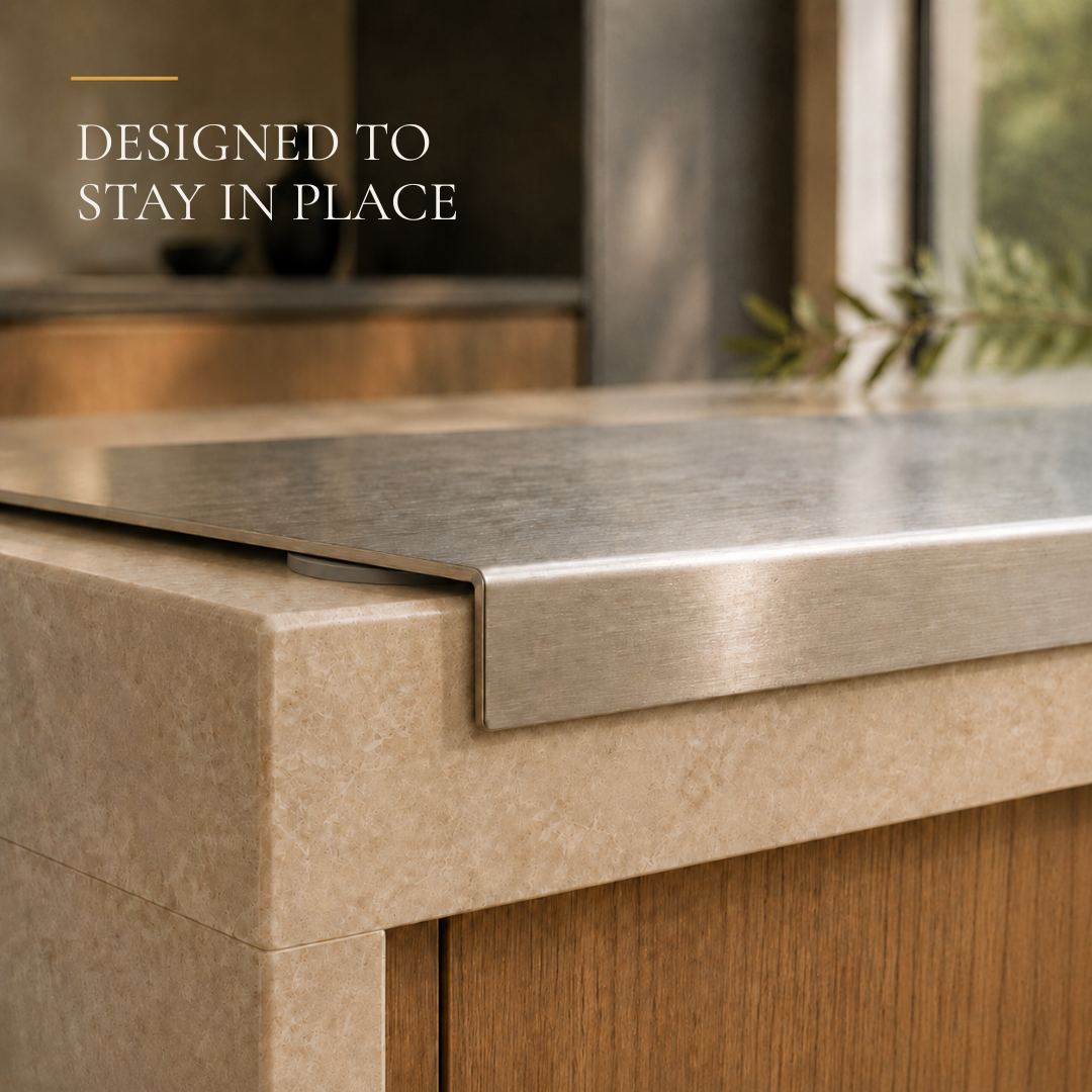 A close-up of a brushed stainless steel countertop with a raised lip over a beige stone slab and wood cabinet, with the text 'DESIGNED TO STAY IN PLACE' in the upper left.
